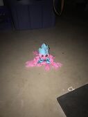 A photo of Phineas Wormthrice in real life. Finny is a doll made of pink yarn sectioned off into five bundles, with black button eyes, a light blue ribbon bowtie, and a matching light blue felt jester cap with golden bells on it. Ey are sitting on the concrete floor of a storage room.